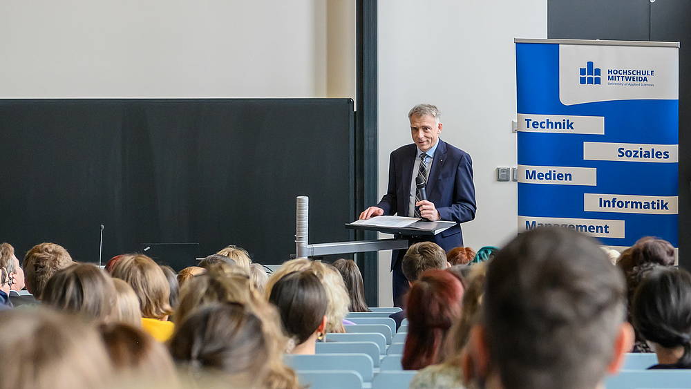 Blick von hinten in den vollbesetzten Hörsaal. Am unteren Ende steht eine männliche Person mit einem Mikrofon in der linken Hand an einem Pult vor dem Rollup, mit dem Logo der Hochschule Mittweida und den Begriffen „Technik“, „Soziales“, „Medien“, Informatik“, Management“ und „Naturwissenschaft“.