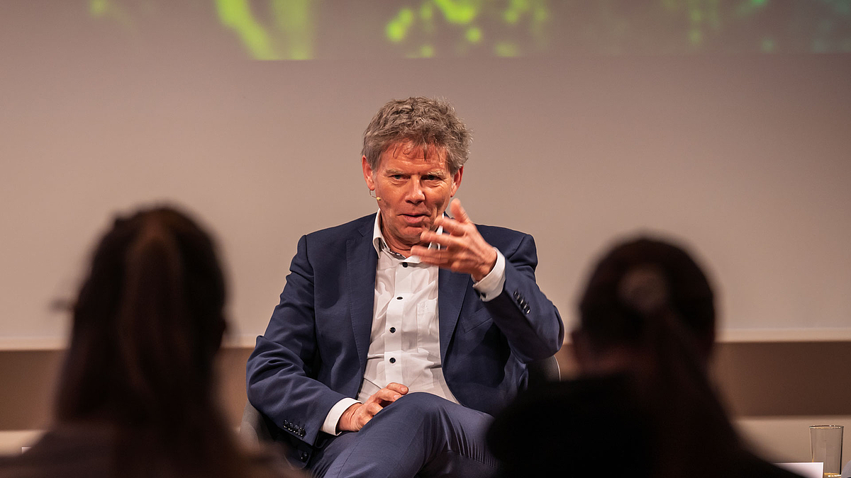 Podiumsteilnehmer sitzend und ernst gestikulierend. Im Vordergrund sich unscharf Köpfe von Zuschauenden zu sehen.