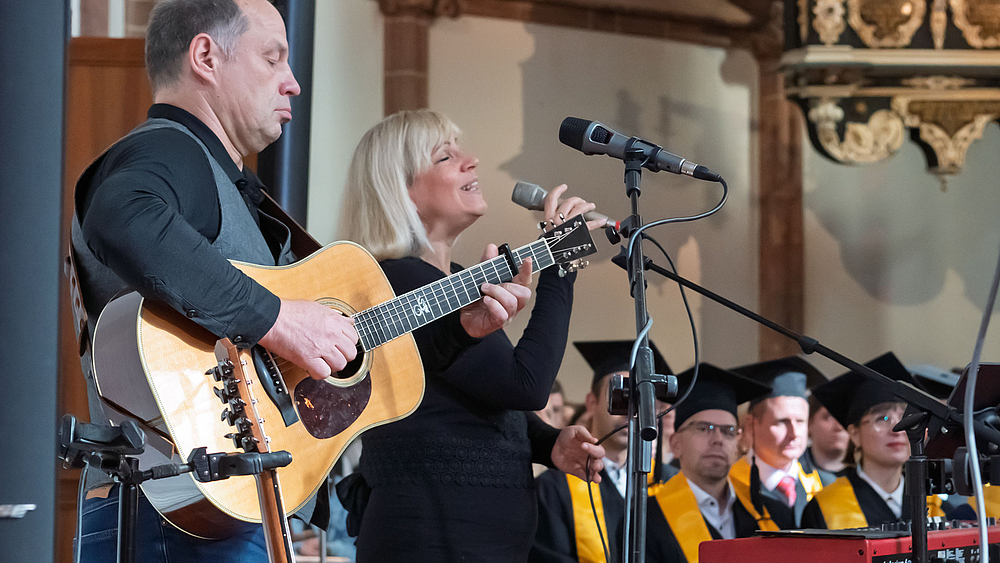 Ein männliche Person mit Gitarre und eine singende weibliche Person am Keyboard mit Mikrofon in der Hand stehen musizieren stehend am Rand des Altarraums, wo Personen mit Doktorhüten zu sehen sind.