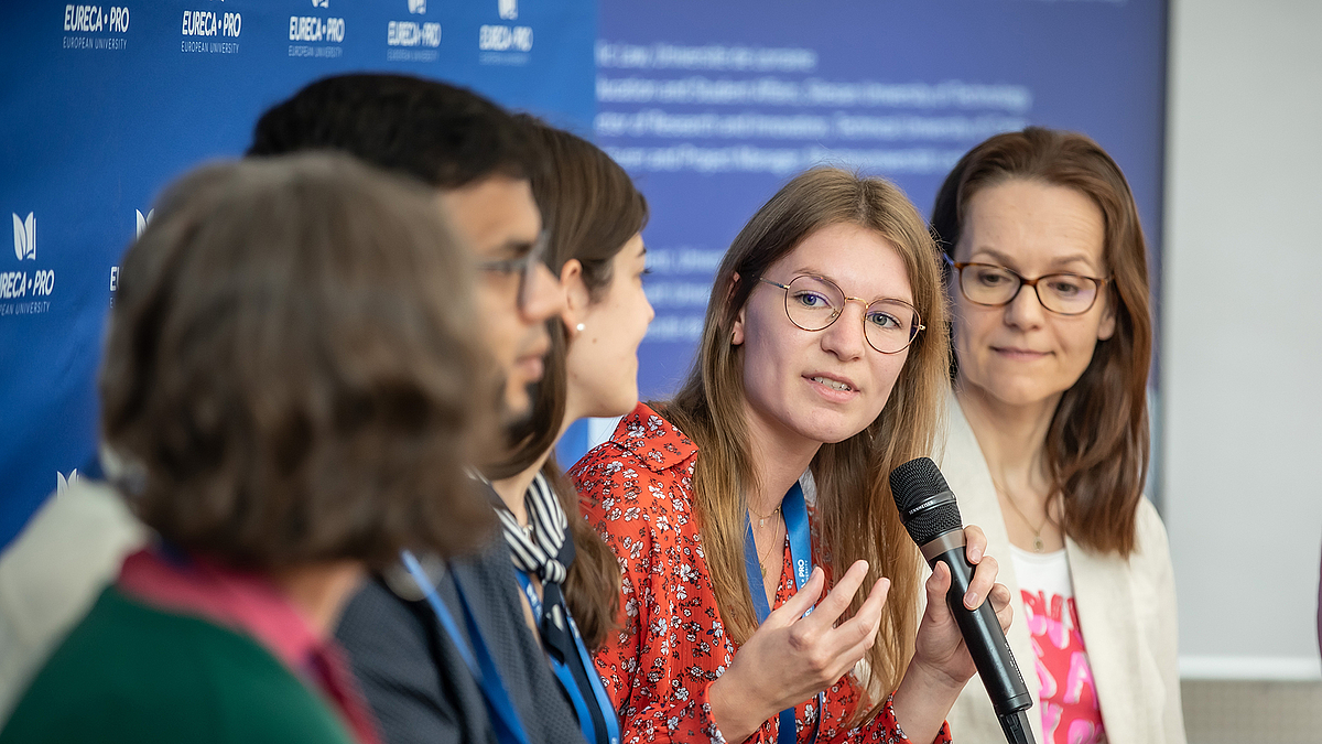 Fünf Menschen sitzen auf einem Panel. Eine junge Frau mit Brille hält das Mikrofon in der Hand und spricht zu den drei links neben ihr Sitzenden.