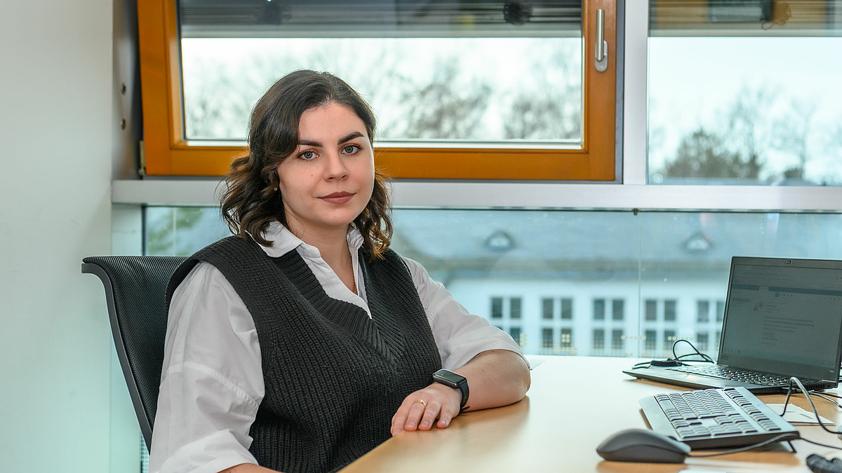 A young woman sits at a desk in front of a window and looks into the camera.