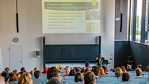 Blick von hinter über die Köpfe der Menschen im Hörsaal nach vorn. Die ältere männliche Person steht am Pult um spricht. Über ihr ist eine Folie projiziert unter anderem mit dem Text: „Pflegende Angehörige. Stille Helden bei der Pflege daheim.“