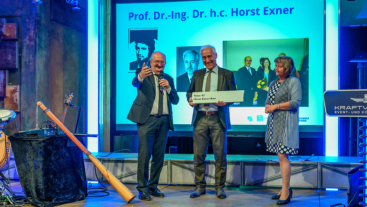 Das Foto zeigt den Rektor der Hochschule, Ludwig Hilmer links und die Kanzlerin rechts auf eine Bühne. Zwischen ihnen steht Horst Exner mit dem ihm soben verliehenen Gebäudeschild mit der Aufschrift "Horst-Exner-Bau".