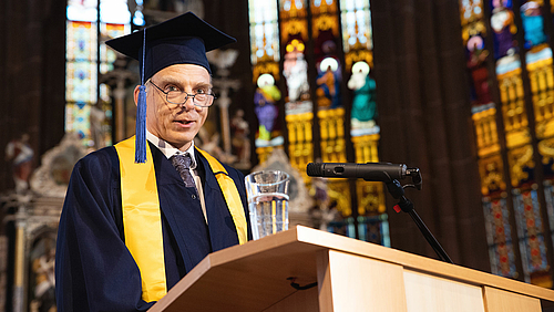 Volker Tolkmitt spricht am Pult im Altarraum der Mittweidaer Stadtkirche. Er trägt einen dunkelblauen Talar und Hut. Im Hintergrund fällt Sonne durch die farbenfrohen Kirchenfenster.