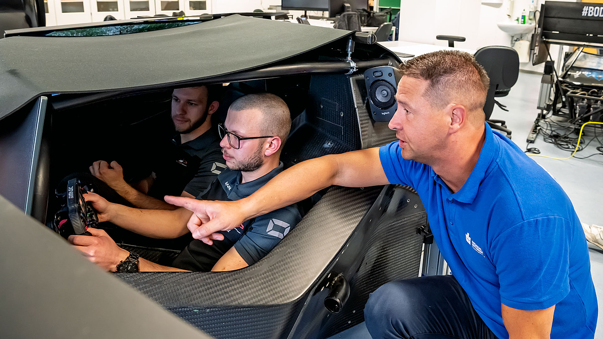 Zwei junge Männer im Cockpit der Simulationsplattform ist. Außen kniet eine weitere männliche Person, die Hinweise gibt.
