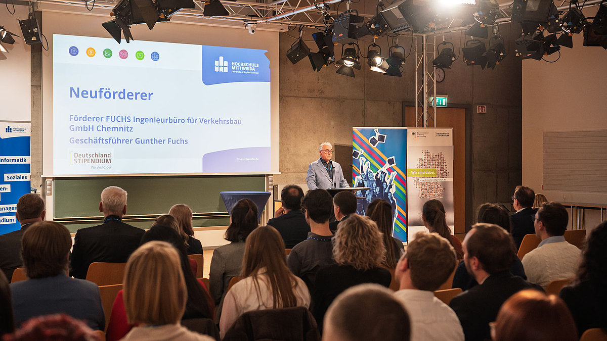 Blick über die Köpfe des Publikums hinweg auf männlichen Redner am Pult. Auf der Leinwand hinter ihm ist unter anderm zu lesen: "Neuförderer. Fuchs Ingenieurbüro für Verkehrsbau GmbH"
