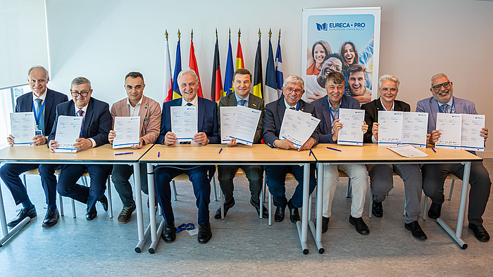 Neun Personen im mittleren Alter sitzen an einem langen Tisch und halten zwei Papierseiten in die Höhe, die sie unterschrieben haben. Im Hintergrund sind die Flaggen aller Länder der EURECA-PRO-Hochschulen und die Europa-Flaggen sichtbar.