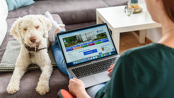 Das Foto zeigt eine weibeliche Person von hinten, die auf ihrem Laptop die Bewerbungsseite der Hochschule Mittweida geöffnet. Der Schriftzug "Jetzt bewerben" ist zu lesen. Auf dem Sofa neben dem Laptop liegt gelassen ein Hund mit weißem Fell.