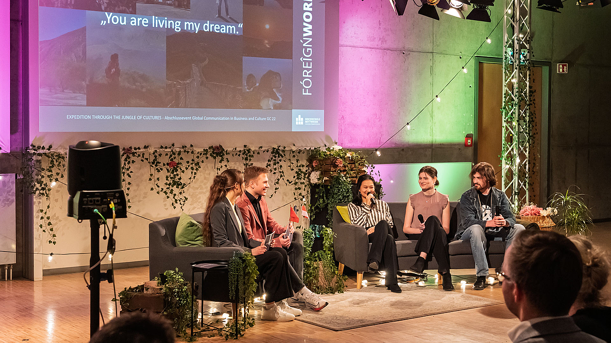 Die beiden Moderator:innen sitzen auf einem Sofa. Auf einem weiteren Sofa rechts von ihnen sitzen zwei junge weibliche Personen und eine junge männliche. Eine der weiblichen Personen spricht in ein Mikrofon. Im Hintergrund sind Reisebilder projiziert. Darüber ist das Zitat „You are living my dream“ gelegt.