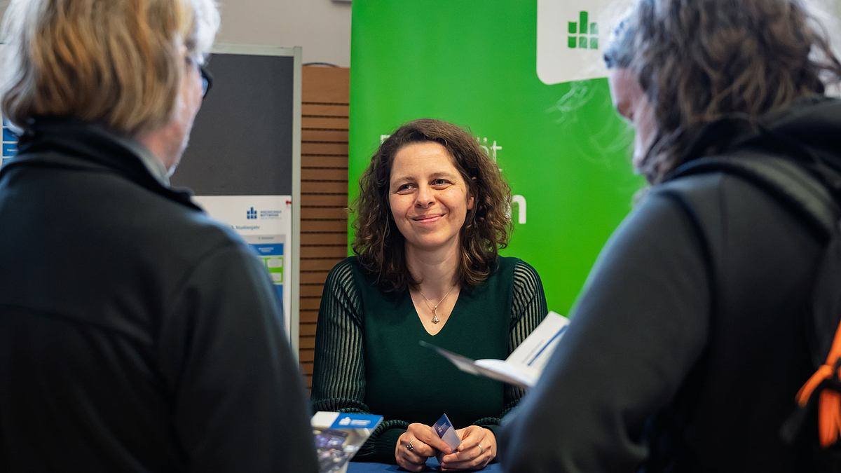Eine lächelnde weibliche Person an einen Tresen vor ienen grünen Rollup unterhält sich mit zwei Personen im Bildvordergrund.