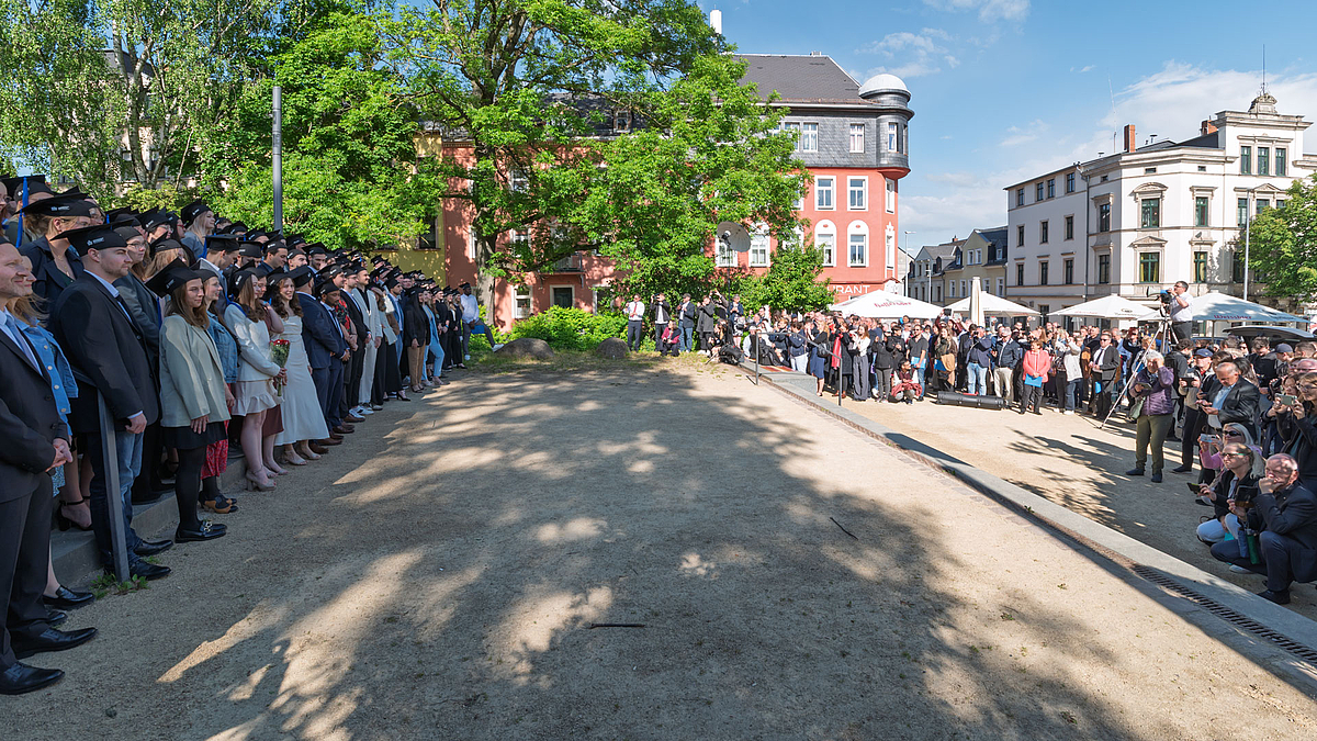 Links die Absolvent:innen auf dem Platz, rechts mit großem Abstand die Angehörigen und der Fotograf des Gruppenfotos auf einer Leiter.