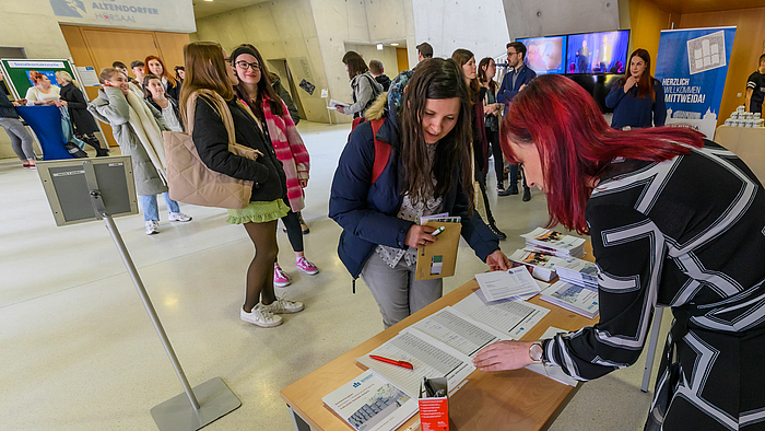 Ein großes Foyer. Einige Menschen stehen in einer Schlange. Die erste Frau in der Schlange steht vor einem Tisch im Vordergrund. Ihre gegenüber auf der anderen Seite des Tisches steht eine weitere weibliche Person. Beide beugen sich über Unterlagen auf dem Tisch.