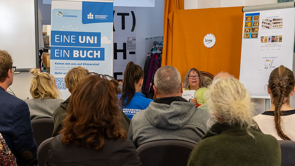 Ein großes Rollup zeigt die Aufschrift "Eine Uni - ein Buch". Im Vordergrund sitzen mehrere Personen in mehreren Sitzreihen auf Stühlen. Im Hintergrund ist eine Frau mit Brille zu erkennen, die ihnen aus einem Buch vorliest.