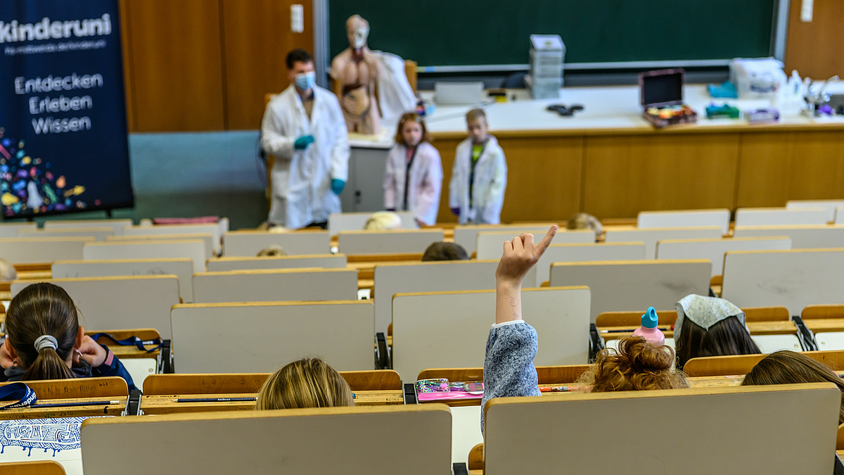 Eine einzelne Hand mit Melde-Finger ragt aus den Sitzreihen des Hörsaals hervor, in denen sonst nur Köpfe von hinten zu sehen sind. Im Bildhintergrund sind unscharf das Pult und die davor stehenden Personen (Dozent, zwei Kinder in weißen Kitteln) sowie der Modelltorso zu erkennen.