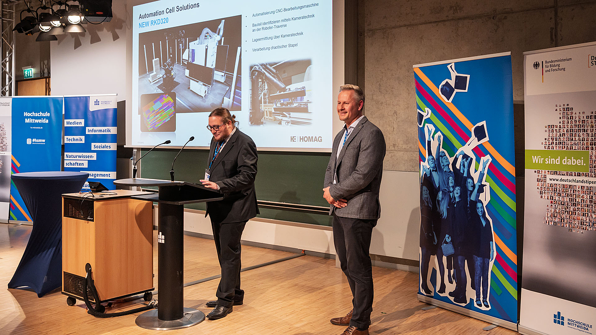 Zwei männliche Personen vor Rollups des Deutschlandstipendiums und einer Folie der HOMAG GmbH auf der Leinwand.
