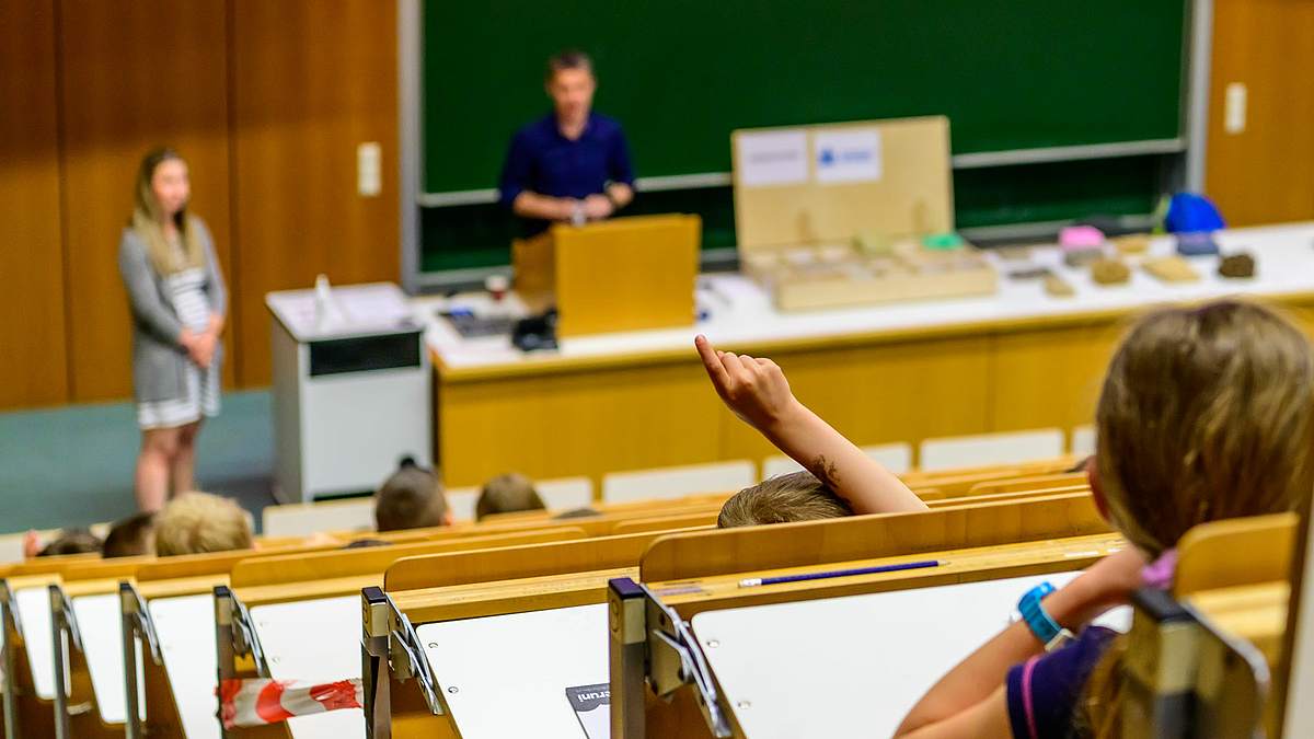 Das Foto zeigt den zum Melden gestreckten Arm eines Kindes vor unscharfen Vorder- und Hintergrund des Hörsaals.