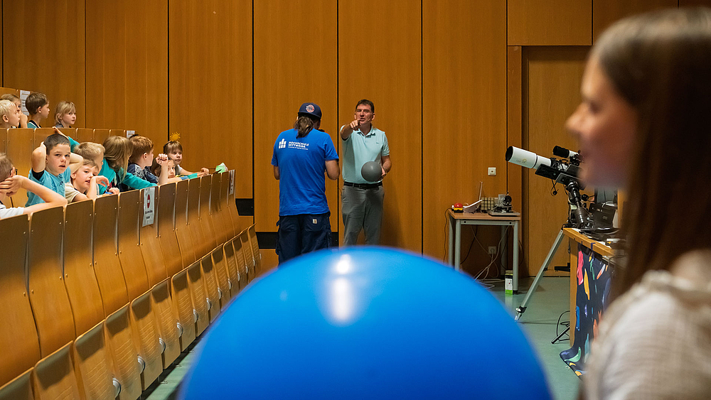 Im Vordergrund steht ein Kind mit einem großen die Erde symboliserenden Ball. im Bilderhintergrund steht ein ein Dozent mit einer kleinen Kugel, dem Mond. Rechts von ihm steht ein Telekop auf einem Stativ.