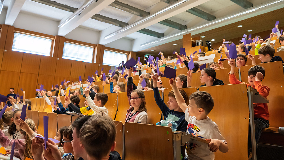 Kinder im Hörsaal halten farbige Karten hoch, hier überwiegend lilafarbene.