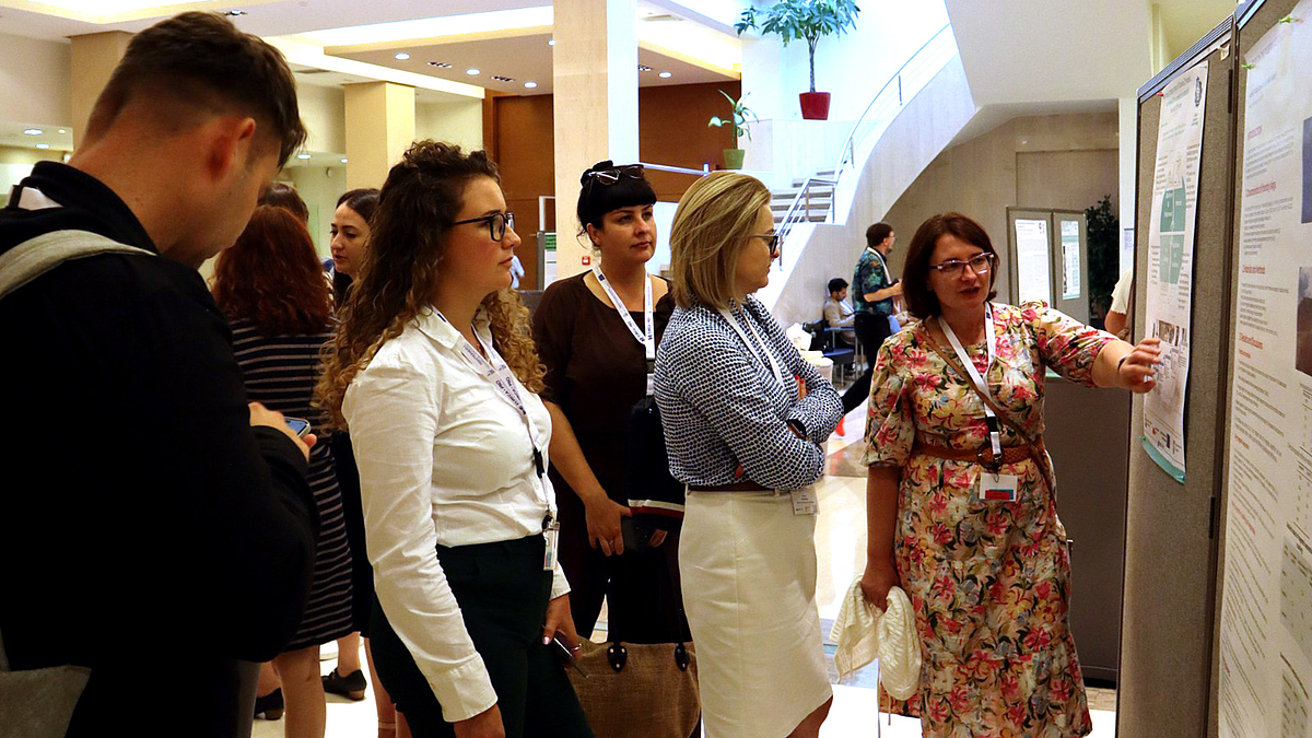 Vier Frauen stehen vor einem wissenschaftlichen Plakat an einer Poster-Tafel. Eine der Frauen erklärt den anderen den Inhalt ihrer Forschung.