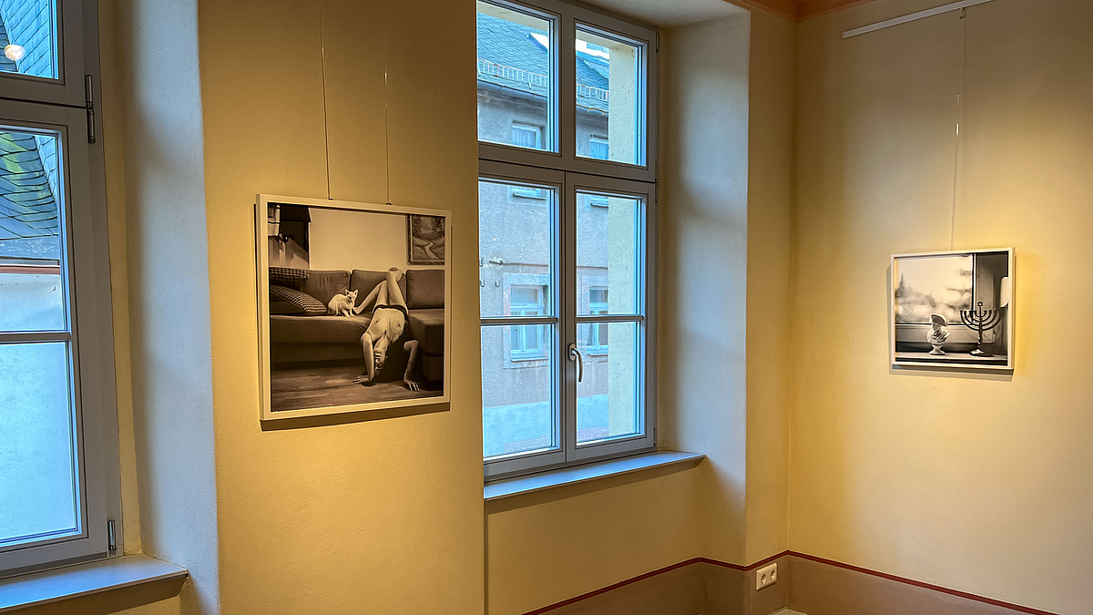 Zwei Fotos in einem Ausstellunsgraum. Das linke zeihgt eine Nackte Frau, die kopfüber von einem Sofa herableht. Neben ishr sitzt eine Katze. Auf dem Foto im rechten Winkeöl zum ersten iszt ien Fenster zusnehen. Davor auf der Fensterbank stehe eine Miniatur-Napleon-Büste und ein kleiner siebenarmiger Leuchter.