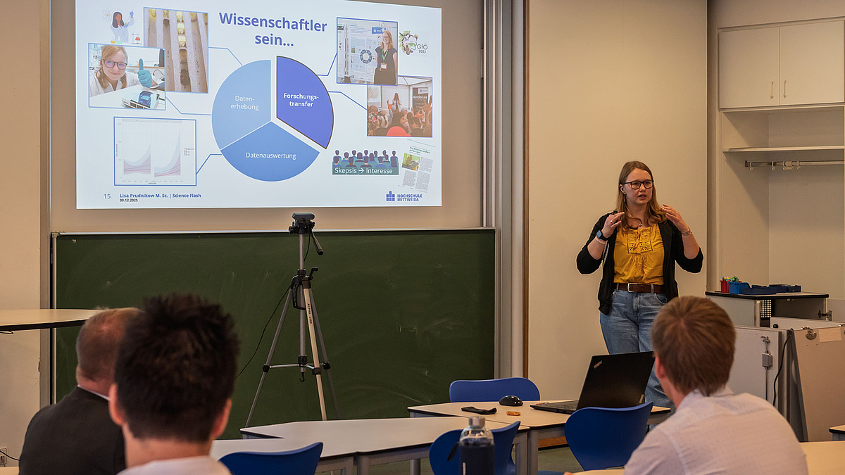 Weibliche Person vor projizierter Powerpointpräsentation. Im Vordergrund Köpfe des Publikums von hinten.