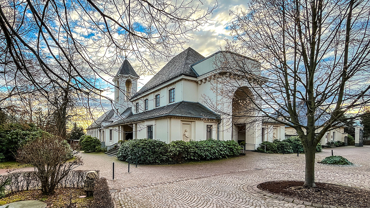 Blick auf ein vor einem in der Abendsonne strahlenden, hellen kirchenartiges Gebäude  mit Turm und großem Engangsportal in einer parkartigen Umgebung mit Bäumen und Sträuchern.