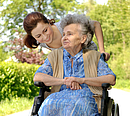 Eine braunhaarige Frau beugt sich freundlich von hinten zu einer alten Frau im Rollstuhl.