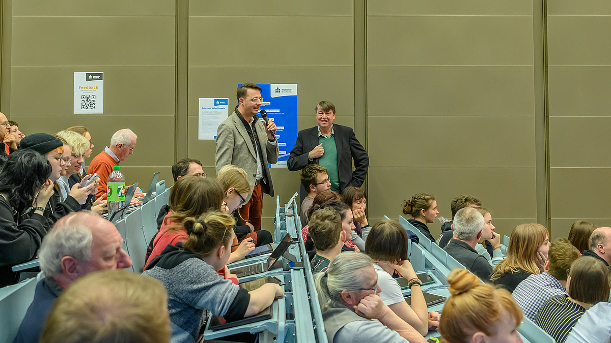 Blick von der Seite durch die von rechts nach links ansteigenden Bankreihen des Hörsaals auf zwei stehende männliche Personen im Bildhintergrund. Die linke Person hält ein Mikrofon in der Hand und spricht in Richtung des nicht sichtbaren Referenten.