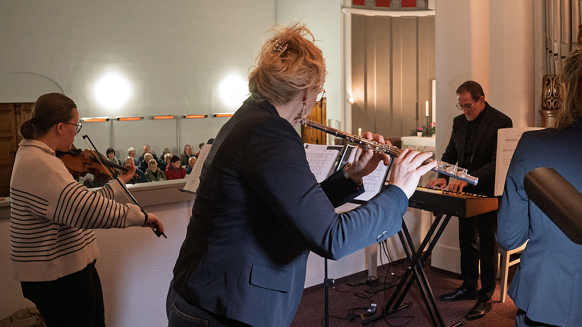 Blick von hinten zwischen den drei Musikerinnen auf den Musiker am Keybor. Im Hintergrund sind Zuhörende zu erkennen.