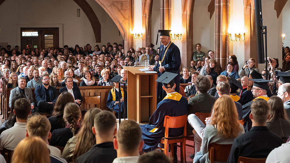 Blick aus dem Altarraum ins Kirchenschiff über die Köpfe der dort sitzenden hinweg. Eine männliche Person im blauen Talar am Rednerpult wendet sich den hinter ihr sitzenden Personen zu.