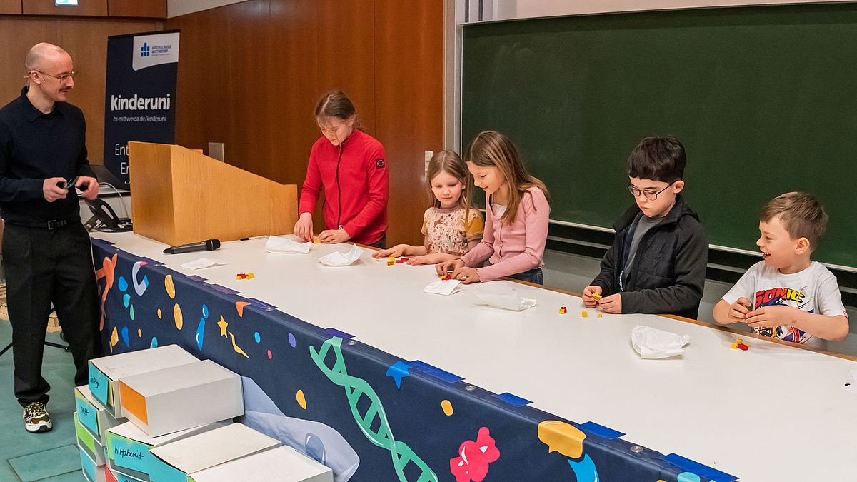 Fünf Kinder stehen nebeneinander hinter dem Hörsaal-Pult und setzen bunte Legosteinen zusammen. Der Dozent schaut den Kindern zu.