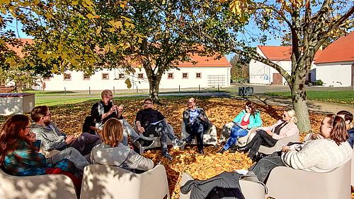 Goldener Oktober im Kloster Nimbschen für den Promotionsworkshop von NextGen, das Qualifikationsprogramm für den professoralen Nachwuchs der Hochschule Mittweida. Das Foto zeigt zehn Personen auf Sesseln im Kreis unter Bäumen sitzend.