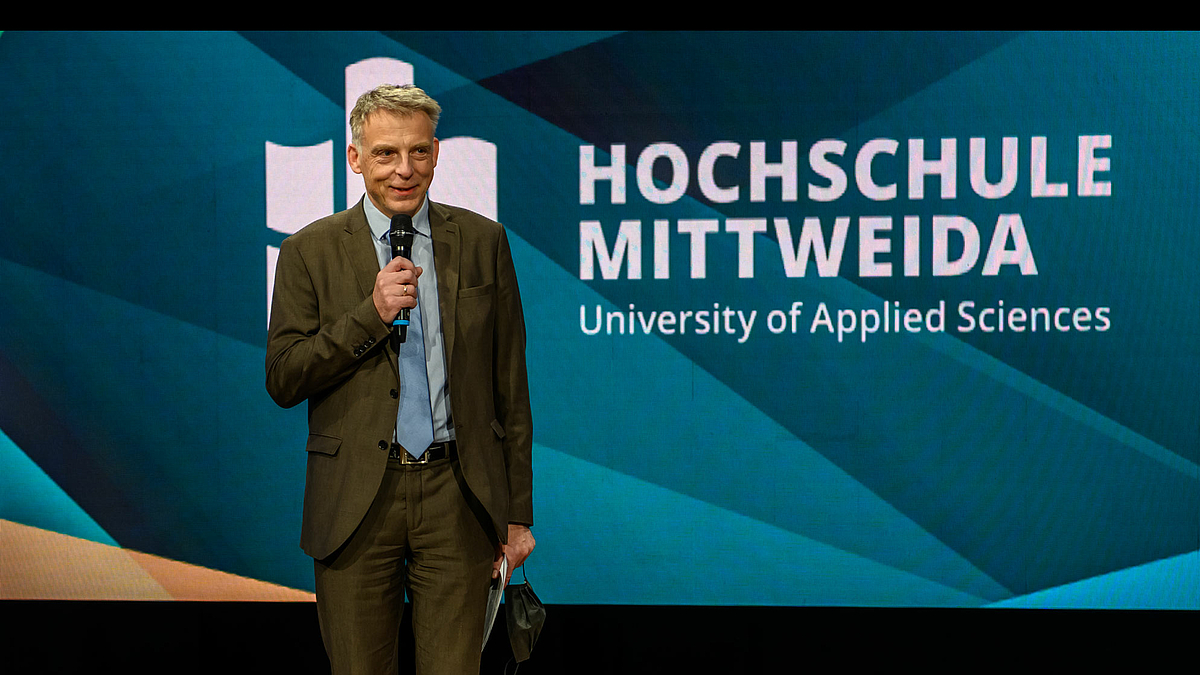 Das Foto zeigt eine lächelnde männliche Person mit einem Mikrofon in der Hand. Der Bildhintergrund ist nahezu vollständig gefüllt mit einem Display, auf dem der Schriftzug Hochschule Mittweida zu lesen ist.