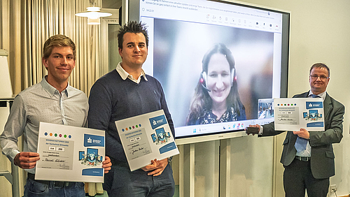 Prorektor Forschung Professor Uwe Mahn (r.) mit den Preisträger:innen des Wissenschaftspreises der HSMW 2025: Pascal Winkler, hier vertreten von Jim Köcher, Justus Reuter und live zugeschaltet Dr. Marika Kaden. Drei männliche Personen mit Urkunden in der Hand. Zwischen ihnen auf einem großen Bildschirm eine weibliche Person.