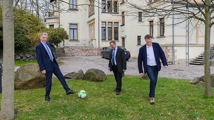 Drei männliche Personen in Anzügen spielen Fußball auf der Wiese vor einem historischen Gebäude, der Direktorenvilla der Hochschule Mittweida.