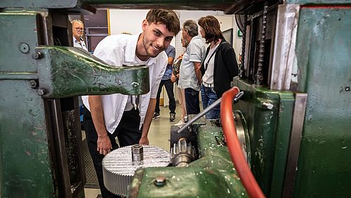 Max Brunner hat den Durchblick – im doppelten Sinn des Wortes. Fast sechs Monate hat der Maschinenbaustudent neben seinem Studium daran gearbeitet, die über siebzig Jahre alte Wälzfräsmaschine flott zu bekommen. Eine junge männliche Person blickt lächelnd durch eine alte Maschine mit Zahnrädern, Wellen und Ketten hindurch. Im Bildhintergrund stehen weitere Personen.