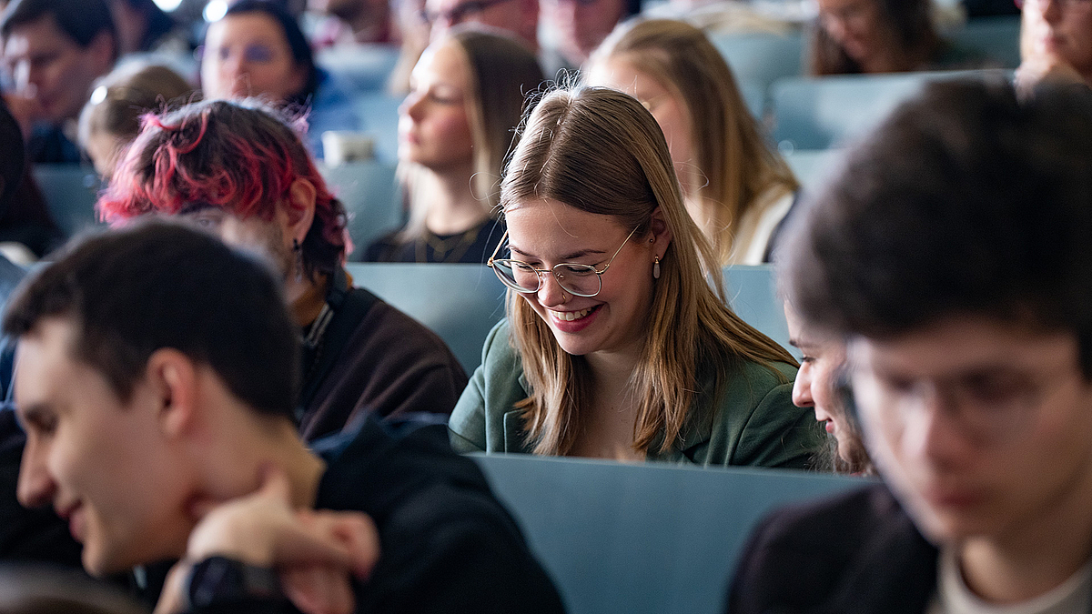 Eine Studentin sitzt im Hörsaal. Sie schaut nach unten und grinst breit mit offenem Mund. Ganz am rechten Bildrand sieht man nur versteckt, dass eine Kommilitonin zu ihr gewandt ist und offenbar mit ihr redet.