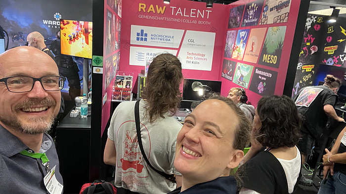 Zwei Menschen schauen freundlich grinsend in die Kamera, während sie ein Selfie machen. Im Hintergrund steht ein junger Mann und spielt an einem Messestand ein Computerspiel.