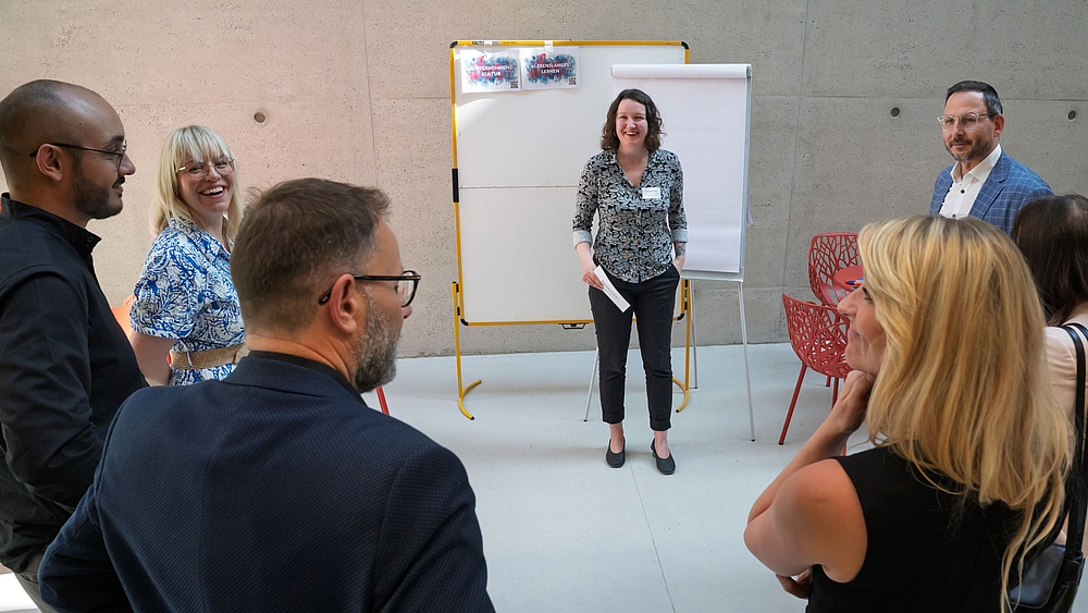 Sechs Personen stehen im Halbkreis, sind teilwese von hinten zusehen. Im Bildhintergrund steht eien weibliche Person lächelnd vor einem Flipchart.