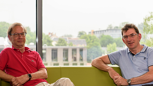Laserkompetenz kombiniert: Prof. Dr. rer. nat. Steffen Weißmantel (links) und Prof. Dr. rer. nat. habil. Alexander Horn leiten die DFG-geförderte Grundlagenforschung im Projekt MULTIPULS. Das Foto zeigt zwei männliche Personen mittleren Alters auf einem Sofa vor einer großen Fensterfläche: Prof. Steffen Weißmantel und Prof. Alexander Horn. Beider Blick ist dem Betrachtenden zugewandt.
