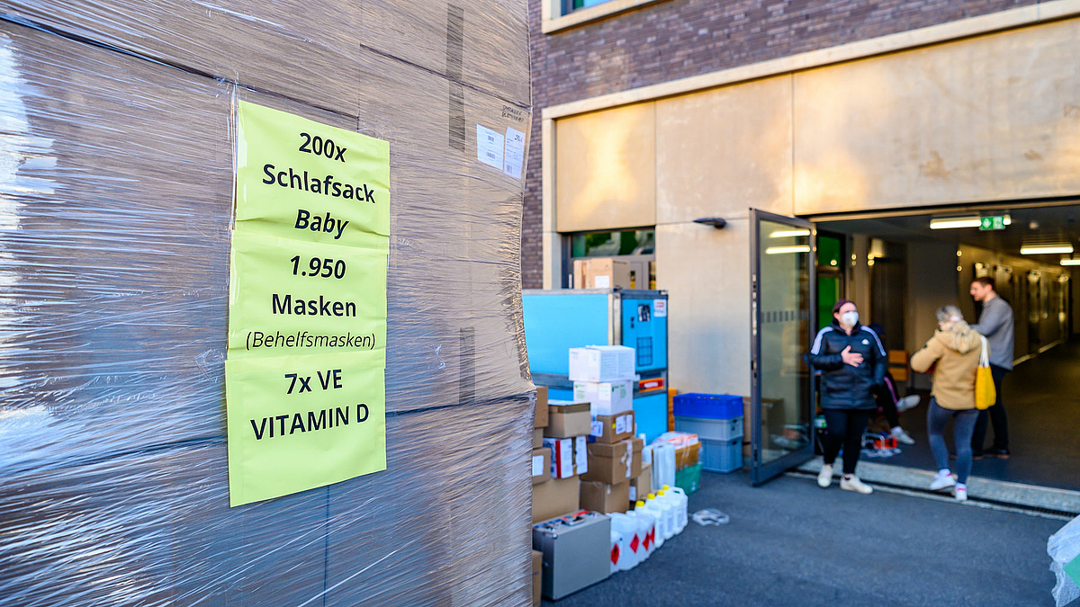 Das Foto zeigt gestapelte Kartons, die mit Folie umwickelt sind. Ein Zettel mit der Aufschrift 200 x Babyschlafsack, 1.950 Behelfsmasken, 7 x VE Vitamin D“ weist auf die Zusammenstellung der Palette hin.