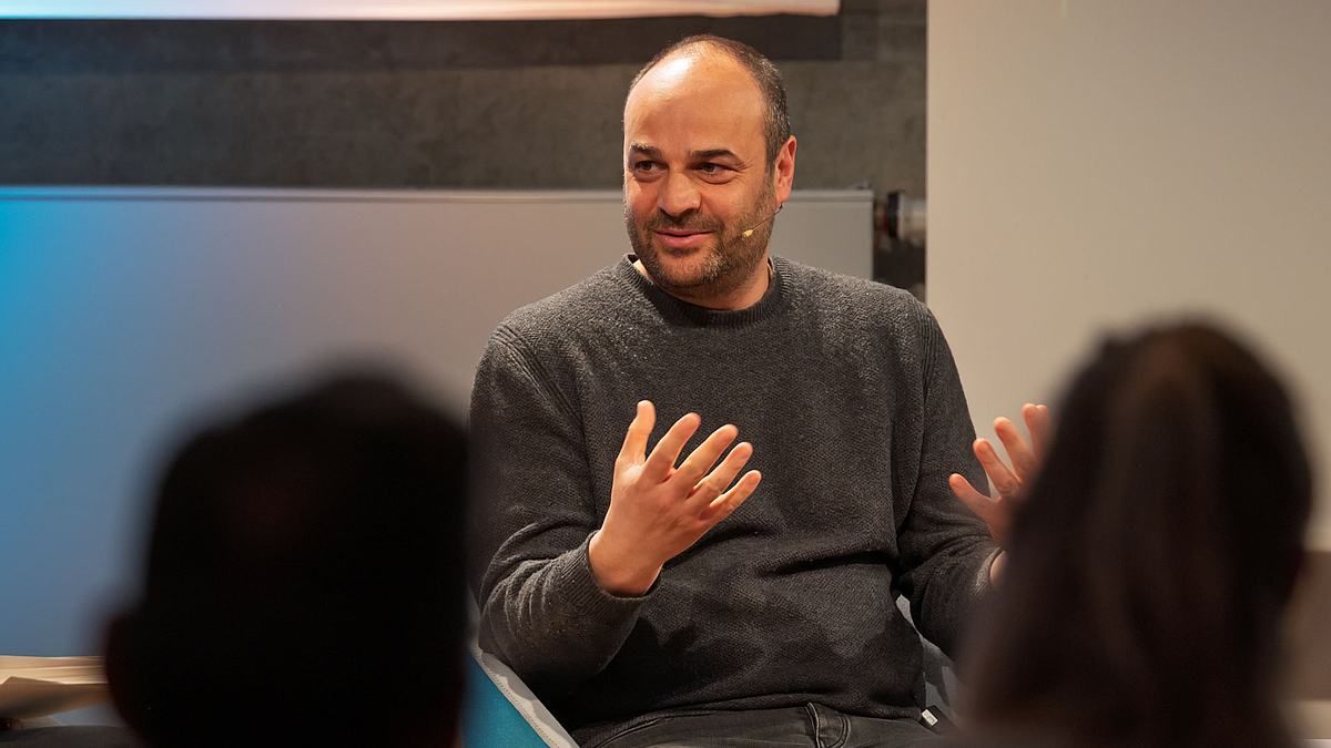 Podiumsteilnehmer sitzend und freundlich gestikulierend. Im Vordergrund sich unscharf Köpfe von Zuschauenden zu sehen.