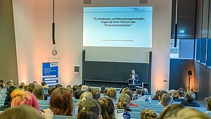 Blick von hinten über die Köpfe der Zuschauenden auf den Dozenten, der vor der Tafel steht. Über ihm zeigt die Projektion den Text: „Zu Pandemien und Bekämpfungsmethoden fragen Sie Ihren Tierarzt oder Tierseuchenbekämpfer.“