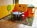 gelbes Spielhaus mit grünem Dach, links daneben steht ein kleiner runder Tisch mit vier bunten Kinderstühlen.