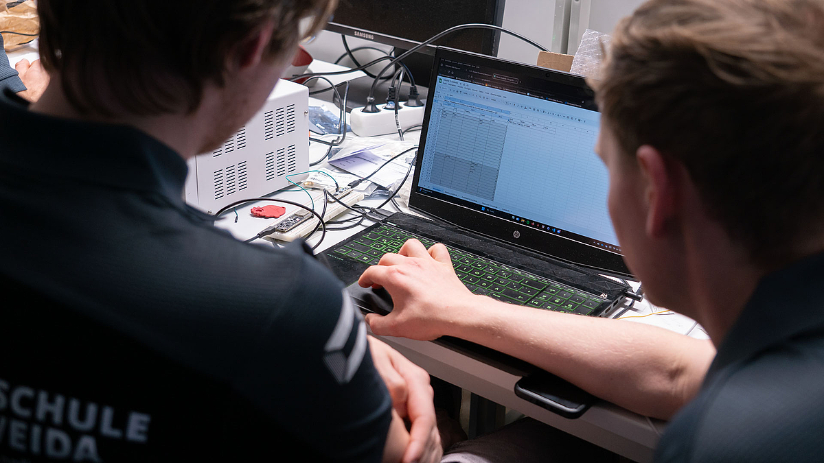 Aufgeklappter Laptop, vor dem zwei männliche Personen sitzen. Auf dem Display ist eine Tabelle mit Werten zu sehen.