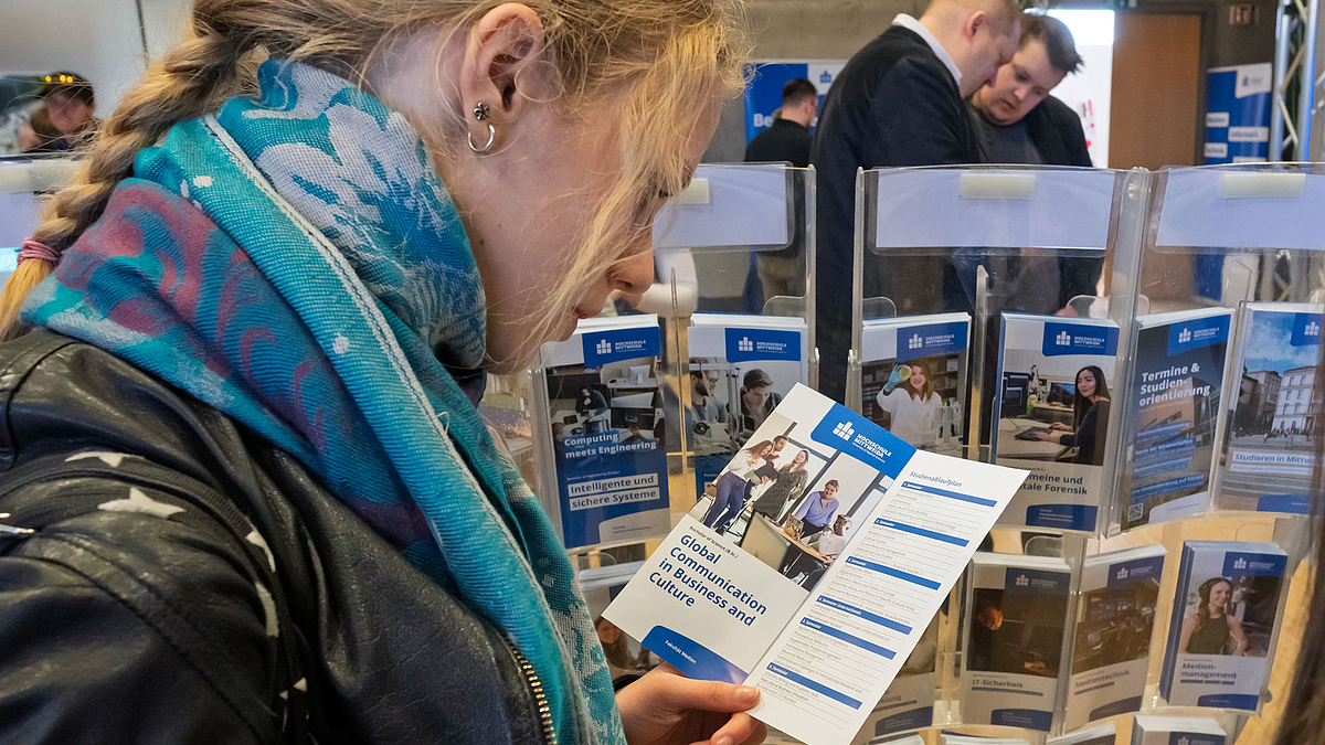 Junge weibliche Person steht vor einem Ständer mit Studiengangsflyern und ist vertieft in den Flyer über "Global Communication in Business and Culture".
