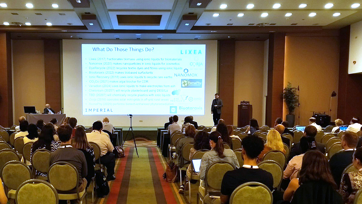 Ein Mann spricht vor einer großen Leinwand auf einem Podium. Die Leinwand zeigt viel Schrift auf weißem Grund. Im Vordergrund sitzen Menschen in circa zehn Sitzreihen im Auditorium.
