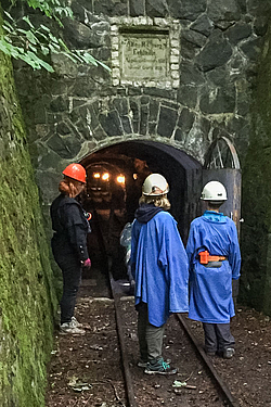 Drei Kinder stehen mit Schutzhelm und Regenjacke vor dem Eingang zu einem Stollen. Bahngleise führen in den Stollen. Die Tür ist geöffnet, gibt den Blick ins Innere frei.