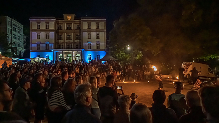 Blick auf das Hauptgbäude der Hochschule bei Naht. Im Vordergrund viele Menschen, die einer Feuershow. 