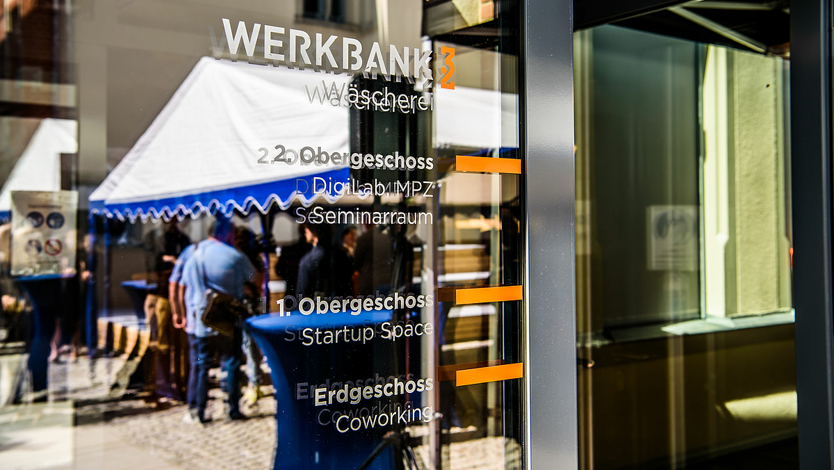 Auf dem Foto ist die gläserne Eingangstür zur ehemaligen Wäscherei zu sehen, auf der die Funktionen der drei Geschosse der Werkbank32 zu lesen sind: „EG: Coworking; 1. OG: Startup Space; 2. OG: DigiLab MPZ, Seminarraum“